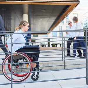 Una ragazza bionda percorre sulla sedia a rotelle una rampa d'accesso all'esterno di un edificio in cui si vedono altre persone camminare