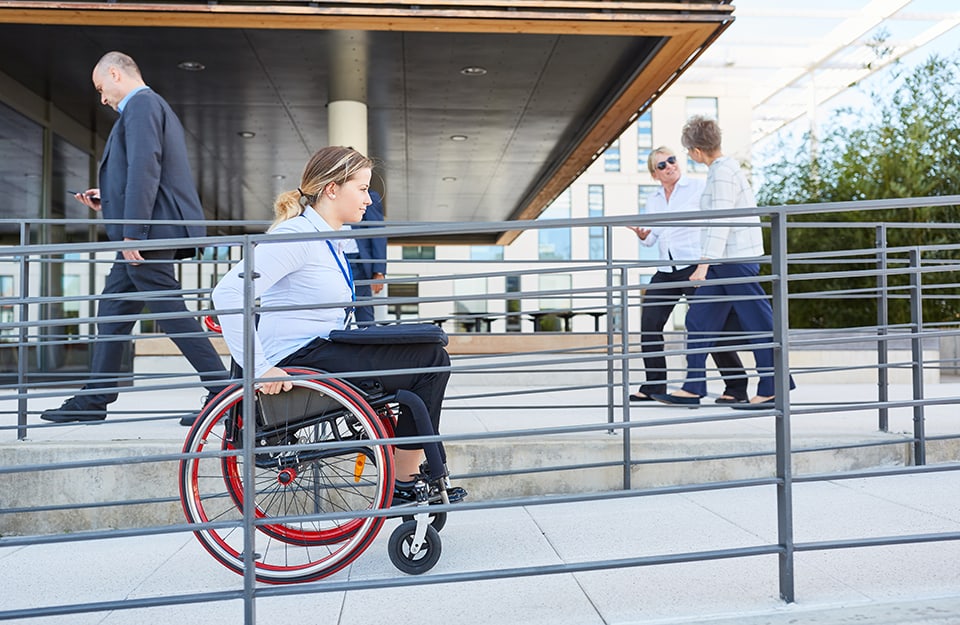 Una ragazza bionda percorre sulla sedia a rotelle una rampa d'accesso all'esterno di un edificio in cui si vedono altre persone camminare