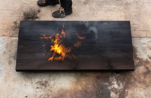 Un uomo che tiene in mano una torcia e brucia un grosso pezzo di quercia creando fiamme e fumo, secondo l'antica tecnica giapponese dello Yakisugi
