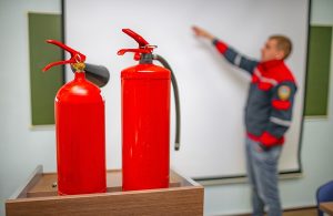 Una lezione sulla sicurezza antincendio, con in primo piano due estintori e sullo sfondo un vigile del fuoco che indica qualcosa su una lavagna