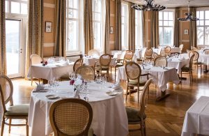 La sala ristorante di un hotel, con tavoli circolari e rettangolari, lunghe tovaglie bianche, sedie in stile classico con schienale impagliato, pavimento in parquet e molte finestre