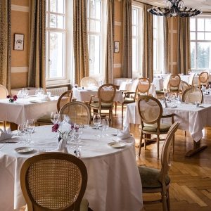 La sala ristorante di un hotel, con tavoli circolari e rettangolari, lunghe tovaglie bianche, sedie in stile classico con schienale impagliato, pavimento in parquet e molte finestre