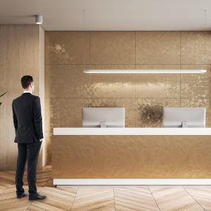 La reception di un hotel sui toni dell'oro e del legno naturale: c'è un addetto alla reception in piedi sul parquet, di spalle, davanti al bancone in cui si vedono due computer