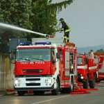 Dei mezzi dei Vigili del Fuoco vicino al recinto in cemento di un'edificio con un pompiere che sta sparando acqua da un cannoncino sul tetto di uno dei camion