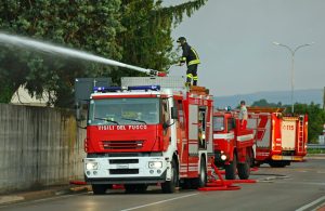Dei mezzi dei Vigili del Fuoco vicino al recinto in cemento di un'edificio con un pompiere che sta sparando acqua da un cannoncino sul tetto di uno dei camion