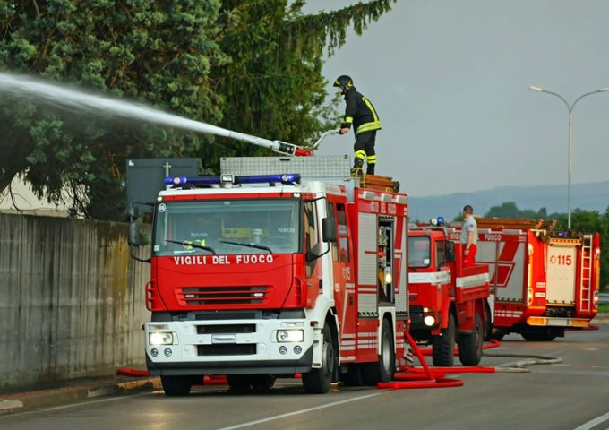 Dei mezzi dei Vigili del Fuoco vicino al recinto in cemento di un'edificio con un pompiere che sta sparando acqua da un cannoncino sul tetto di uno dei camion