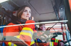 Due Vigili del Fuoco, un uomo e una donna, nell'abitacolo di un'autopompa, con lui che guida e lei che parla via radio