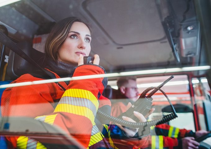 Due Vigili del Fuoco, un uomo e una donna, nell'abitacolo di un'autopompa, con lui che guida e lei che parla via radio