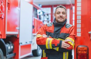 Un veterano dei Vigili del Fuoco a braccia conserte e sorridente all'interno di una caserma, accanto a un'autopompa