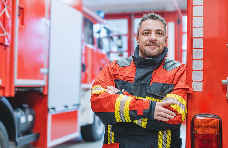 Un veterano dei Vigili del Fuoco a braccia conserte e sorridente all'interno di una caserma, accanto a un'autopompa
