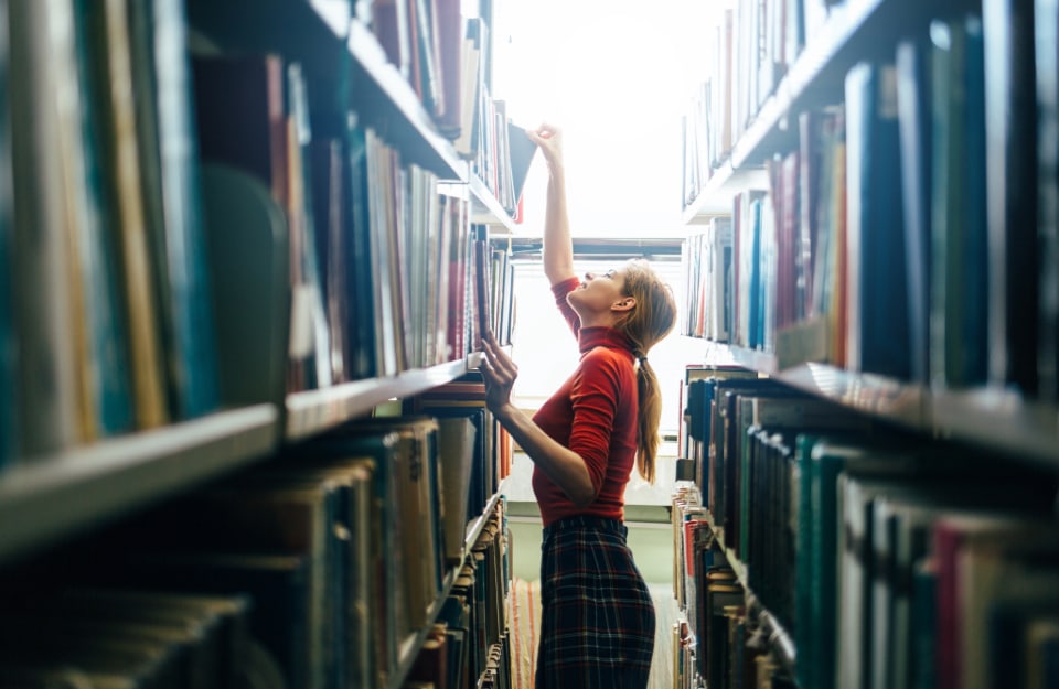 Una giovane bibliotecaria con golfino rosso e gonna a scacchi sta prendendo un libro sui ripiani alti di uno scaffale nello stretto corridoio di una biblioteca