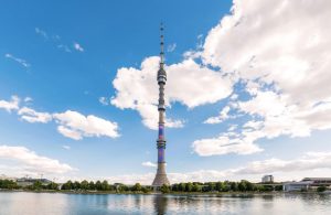 Vista da lontano della torre di Ostankino, a Mosca, che si staglia sul panorama cittadino e si riflette sull'acqua in una giornata col cielo azzurro e alcune nuvole bianche
