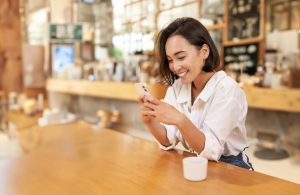 Una ragazza orientale sorride mentre guarda il suo smartphone seduta su un lungo tavolo in una caffetteria in stile anglosassone con arredi in legno