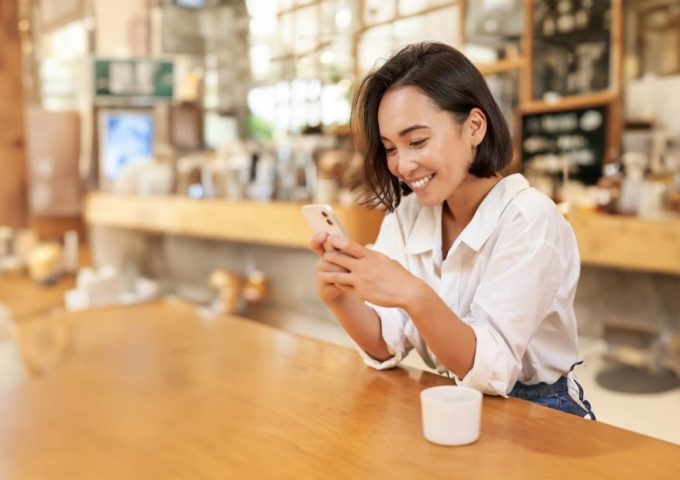 Una ragazza orientale sorride mentre guarda il suo smartphone seduta su un lungo tavolo in una caffetteria in stile anglosassone con arredi in legno