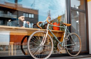 Vista dall'esterno di una vetrina di un locale, con una bicicletta da corsa appoggiata sulla vetrina all'esterno e, dentro, un ragazzo con la barba lunga e lo stile hipster che sta seduto a un tavolino