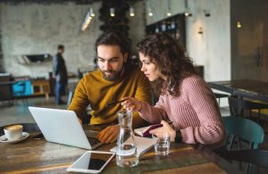 Un ragazzo con la barba e una ragazza stanno discutendo di lavoro davanti a un computer portatile al tavolino di una caffetteria