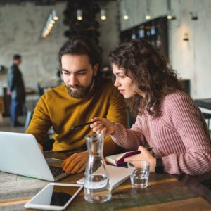 Un ragazzo con la barba e una ragazza stanno discutendo di lavoro davanti a un computer portatile al tavolino di una caffetteria