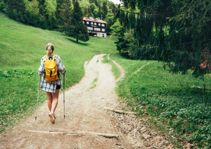 Una ragazza bionda in pantaloncini, vista di spalle, sta camminando con i bastoni da trekking lungo un sentiero che porta a un rifugio alpino tra i boschi