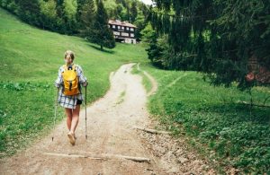 Una ragazza bionda in pantaloncini, vista di spalle, sta camminando con i bastoni da trekking lungo un sentiero che porta a un rifugio alpino tra i boschi