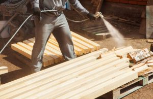 Un uomo, visto dal busto in giù, sta trattando delle travi di legno con della vernice ignifuga applicata a spruzzo