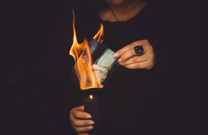 Una donna di cui non si vede il volto, vestita di nero su sfondo nero, sta bruciando una fotografia in un barattolo scuro