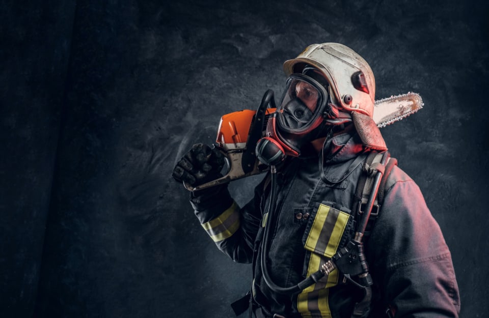 Un pompiere con casco e maschera fotografato di profilo con una motosega poggiata sulla spalla su un sfondo monocromatico scuro