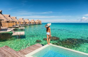 Una donna in costume con un grande cappello, vista di spalle in piedi sul bordo di una piscina di un villaggio turistico, con l'acqua cristallina del mare a pochi passi, e una fila di bungalow su palafitte