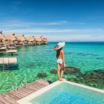 Una donna in costume con un grande cappello, vista di spalle in piedi sul bordo di una piscina di un villaggio turistico, con l'acqua cristallina del mare a pochi passi, e una fila di bungalow su palafitte