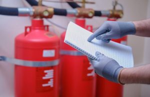 Un ispettore della sicurezza antincendio sta controllando i suoi appunto in un impianto industriale: si vedono le mani dell'uomo, che tengono un blocco con gli appunti, accanto a degli estintori