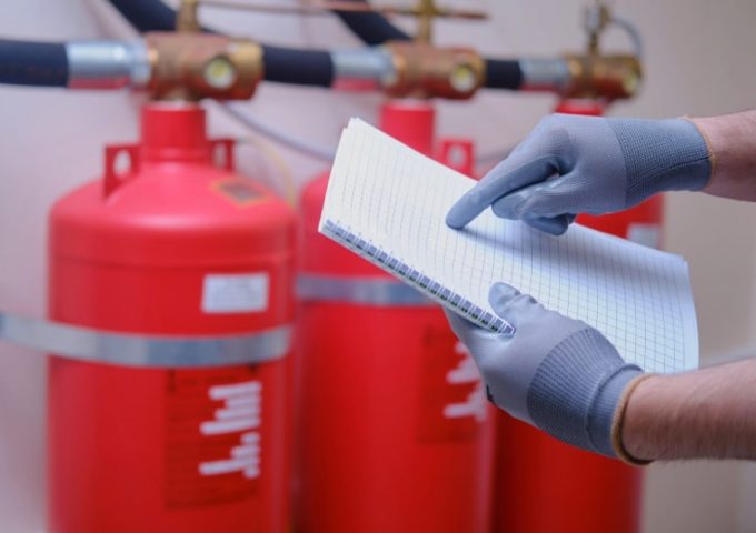 Un ispettore della sicurezza antincendio sta controllando i suoi appunto in un impianto industriale: si vedono le mani dell'uomo, che tengono un blocco con gli appunti, accanto a degli estintori