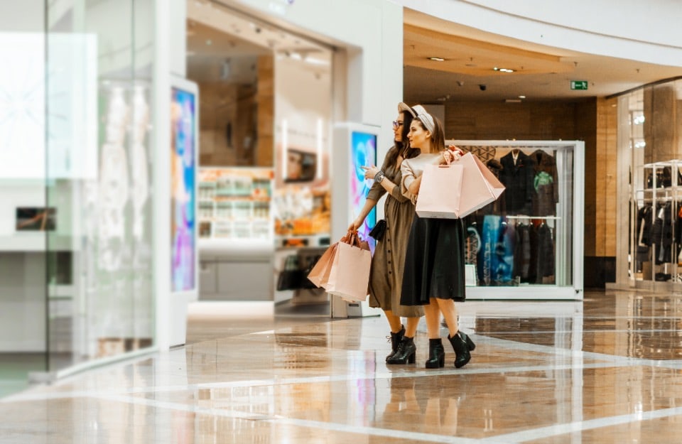 Due ragazze osservano una vetrina di un centro commerciale con delle buste di acquisti in mano