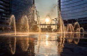 Vista di Porta Garibaldi, a Milano, di tardo pomeriggio, con il sole calante che crea effetti visivi sugli spruzzi d'acqua di una fontana