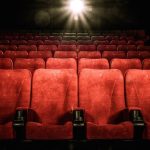 Una sala cinematografica vuota con file di sedili rossi. I sedili sono numerati. La luce del proiettore è accesa, illuminando la parte centrale della sala
