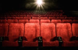 Una sala cinematografica vuota con file di sedili rossi. I sedili sono numerati. La luce del proiettore è accesa, illuminando la parte centrale della sala