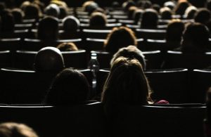 Sala cinematografica affollata con sedili e persone viste da dietro, poco illuminate dalla luce delle schermo (che non si vede)