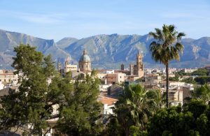 Veduta panoramica di Palermo. In primo piano, ci sono diversi alberi, tra cui una palma alta e alcuni pini. Sullo sfondo, si possono vedere vari edifici storici, tra cui la Cattedrale di Palermo con la sua cupola verde e le torri campanarie. Dietro la città, si ergono le montagne che circondano Palermo, con un cielo sereno, con poche nuvole