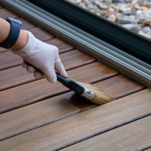 Una mano che indossa un guanto di plastica bianco mentre utilizza un pennello per verniciare o trattare una superficie di legno. La superficie di legno è composta da assi parallele e la parte già trattata appare più scura e lucida rispetto alla parte ancora non trattata, che è più chiara e opaca. Sullo sfondo, si possono vedere dei ciottoli dietro una finestra o una porta scorrevole