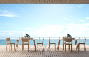 La terrazza di un ristorante con vista sul mare. La terrazza ha un pavimento in legno ed è arredata con tavoli e sedie in legno chiaro, ognuno decorato con piante in vasi bianchi