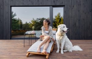 Una ragazza è seduta su una sdraio in legno sulla terrazza, pure in legno, del bungalow di un resort. Accanto alla ragazza c'è un grande cane bianco che guarda in direzione opposta a quella della donna. Il bungalow è in legno scuro con una grande porta-finestra scorrevole