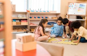 Classe di scuola materna con quattro bambini seduti su un tappeto verde chiaro, impegnati in un'attività con dei blocchi colorati. Sullo sfondo, ci sono scaffali con vari materiali didattici, tra cui forme geometriche, libri e cesti. A destra, c'è una lavagna con dei disegni colorati. In primo piano, sfocato, c'è un tavolo rosso con dei blocchi di legno numerati