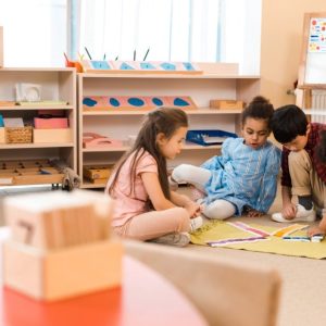Classe di scuola materna con quattro bambini seduti su un tappeto verde chiaro, impegnati in un'attività con dei blocchi colorati. Sullo sfondo, ci sono scaffali con vari materiali didattici, tra cui forme geometriche, libri e cesti. A destra, c'è una lavagna con dei disegni colorati. In primo piano, sfocato, c'è un tavolo rosso con dei blocchi di legno numerati