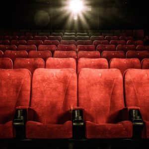 Una sala cinematografica vuota con file di sedili rossi. I sedili sono numerati. La luce del proiettore è accesa, illuminando la parte centrale della sala
