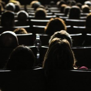 Sala cinematografica affollata con sedili e persone viste da dietro, poco illuminate dalla luce delle schermo (che non si vede)