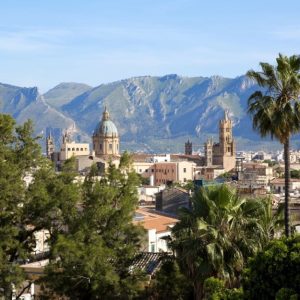 Veduta panoramica di Palermo. In primo piano, ci sono diversi alberi, tra cui una palma alta e alcuni pini. Sullo sfondo, si possono vedere vari edifici storici, tra cui la Cattedrale di Palermo con la sua cupola verde e le torri campanarie. Dietro la città, si ergono le montagne che circondano Palermo, con un cielo sereno, con poche nuvole