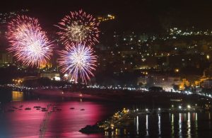 Spettacoli pirotecnici notturni a Palermo durante la festa di Sant'Agata. In primo piano, ci sono tre grandi fuochi d'artificio che esplodono nel cielo, illuminando la città sottostante con colori vivaci come il rosso, il blu e il bianco. Sullo sfondo, si vede la città di Palermo con le sue luci notturne che brillano, creando un'atmosfera festosa. La costa è visibile con diverse barche ancorate nell'acqua, che riflette i colori dei fuochi d'artificio