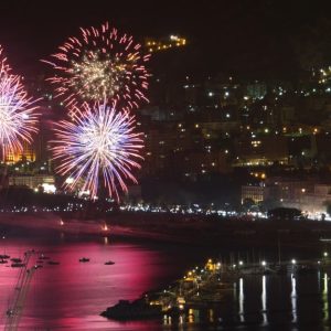 Spettacoli pirotecnici notturni a Palermo durante la festa di Sant'Agata. In primo piano, ci sono tre grandi fuochi d'artificio che esplodono nel cielo, illuminando la città sottostante con colori vivaci come il rosso, il blu e il bianco. Sullo sfondo, si vede la città di Palermo con le sue luci notturne che brillano, creando un'atmosfera festosa. La costa è visibile con diverse barche ancorate nell'acqua, che riflette i colori dei fuochi d'artificio