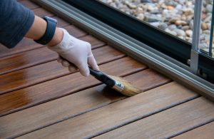 Una mano che indossa un guanto di plastica bianco mentre utilizza un pennello per verniciare o trattare una superficie di legno. La superficie di legno è composta da assi parallele e la parte già trattata appare più scura e lucida rispetto alla parte ancora non trattata, che è più chiara e opaca. Sullo sfondo, si possono vedere dei ciottoli dietro una finestra o una porta scorrevole