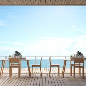 La terrazza di un ristorante con vista sul mare. La terrazza ha un pavimento in legno ed è arredata con tavoli e sedie in legno chiaro, ognuno decorato con piante in vasi bianchi