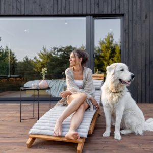 Una ragazza è seduta su una sdraio in legno sulla terrazza, pure in legno, del bungalow di un resort. Accanto alla ragazza c'è un grande cane bianco che guarda in direzione opposta a quella della donna. Il bungalow è in legno scuro con una grande porta-finestra scorrevole