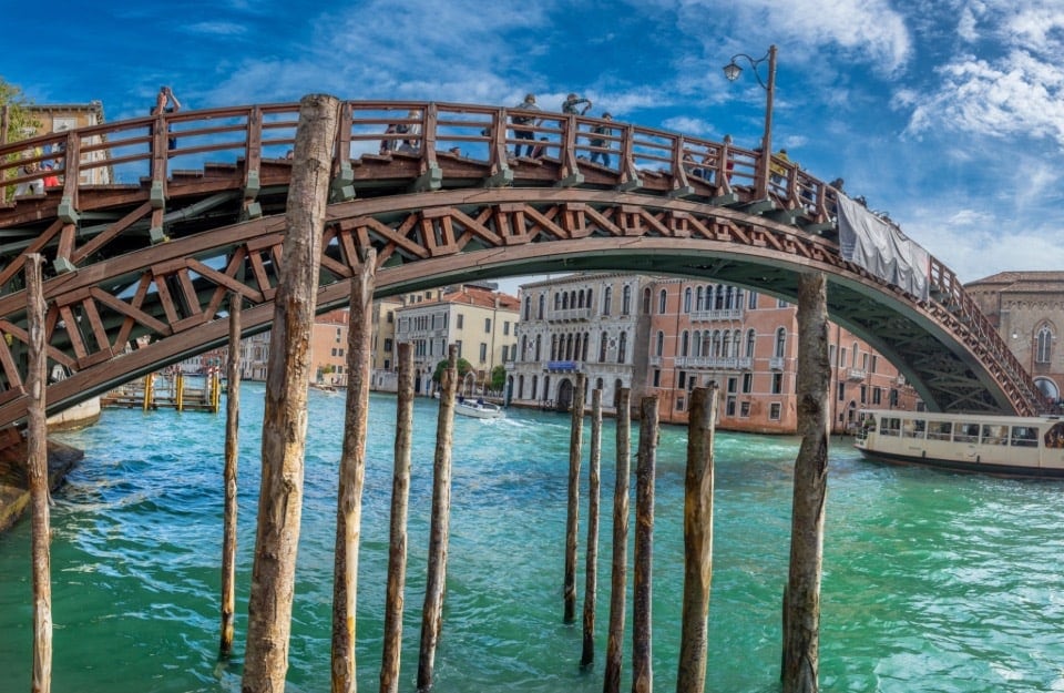 Un ponte di legno che attraversa un canale a Venezia, Italia. Il ponte è costruito con una struttura a traliccio di travi in legno e ha una ringhiera sulla parte superiore. Sotto il ponte, ci sono diversi pali di legno che sporgono dall'acqua. L'acqua del canale è di un vibrante colore turchese e ci sono edifici con la classica architettura veneziana che fiancheggiano il canale. Una barca è visibile sul lato destro dell'immagine e il cielo è sereno con alcune nuvole sparse